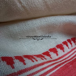 Pode incluir: Close-up de um tecido creme com uma borda estampada em vermelho. O tecido tem uma textura tecida e um design decorativo ao longo de uma borda. O texto "vinayaktextileindia" &eacute; vis&iacute;vel.
