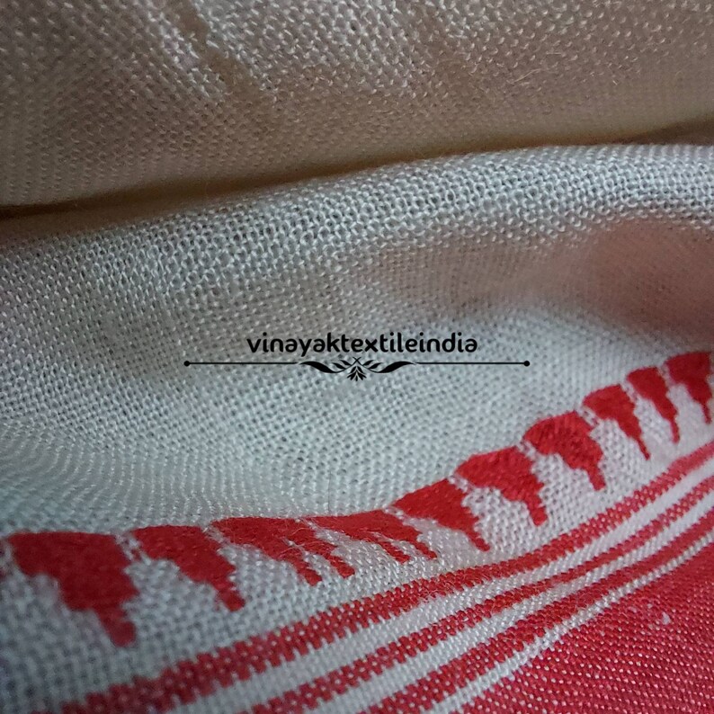 Pode incluir: Close-up de um tecido creme com uma borda estampada em vermelho. O tecido tem uma textura tecida e um design decorativo ao longo de uma borda. O texto "vinayaktextileindia" &eacute; vis&iacute;vel.