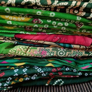 May include: A stack of folded fabric featuring various patterns including green, yellow, and red geometric shapes, small white dots, and intricate block prints. Some layers display metallic embellishments and sequins on the textile surface.