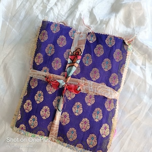 May include: A stack of purple fabric squares with a gold and turquoise pattern. The fabric is tied together with a white and pink ribbon.