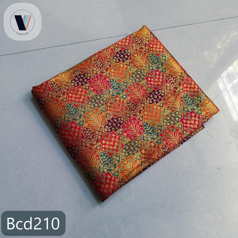 Puede incluir: Una pieza de tela cuadrada con un patr&oacute;n geom&eacute;trico repetitivo de estrellas y motivos de hojas en tonos rojo, naranja, dorado, morado y verde azulado. El tejido tiene un brillo met&aacute;lico y lleva el texto Bcd210 en la esquina.