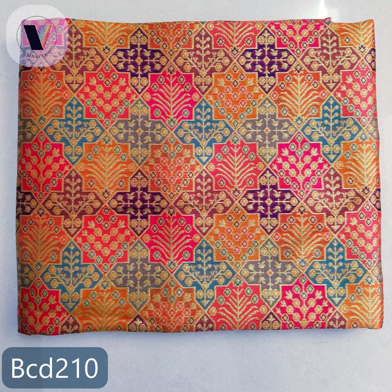 Puede incluir: Una pieza de tela cuadrada con un patr&oacute;n geom&eacute;trico repetitivo de rombos y cruces en tonos vibrantes de naranja, rosa, morado y azul, con intrincados motivos florales dorados tejidos en todo el dise&ntilde;o.