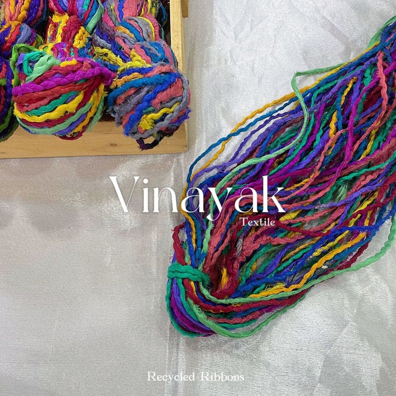 May include: A close-up of a bundle of colourful recycled fabric ribbons. The ribbons are made from scraps of fabric and are tied together in a loose knot. The ribbons are a variety of colours, including red, blue, green, yellow, and purple. The text "Vinayak Textile" and "Recycled Ribbons" is visible in the image.