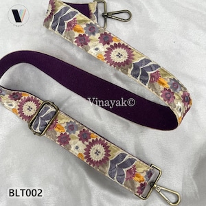 Op de afbeelding: Twee geborduurde tasriemen met bloempatronen in paars, geel en crème. De riemen hebben metalen hardware en een effen paarse achterkant. De tekst "Vinayak©" en "BLT002" zijn zichtbaar.