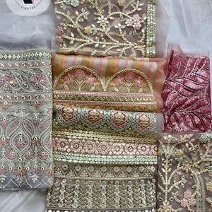 Könnte beinhalten: Eine Sammlung von Stoffmustern mit aufwendigen Stickereien, Metallfäden, Pailletten und Spiegelarbeiten. Die Textilien zeigen verschiedene florale und geometrische Muster in Rosa-, Gold-, Grün- und Grautönen auf einem transparenten Netzstoff.