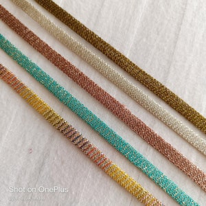 May include: Five different metallic trims with a variety of colors and textures. The trims are arranged in a diagonal line on a white background.  Shot on OnePlus. By vinayak textile.
