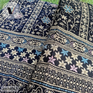 Puede incluir: Tela negra con bordado dorado y azul. La tela tiene un patrón repetitivo de flores y estrellas. La tela es suave y lujosa.