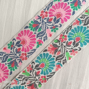 Peut inclure: Deux galons en tissu blanc avec des broderies florales colorées. Le galon du haut a des fleurs roses, rouges, vertes et bleues. Le galon du bas a des fleurs turquoise, rouges, vertes et noires.