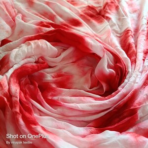 May include: A close-up of a white and red tie-dye fabric. The fabric is folded and draped, creating a swirling pattern. The fabric is soft and flowing, with a subtle sheen.