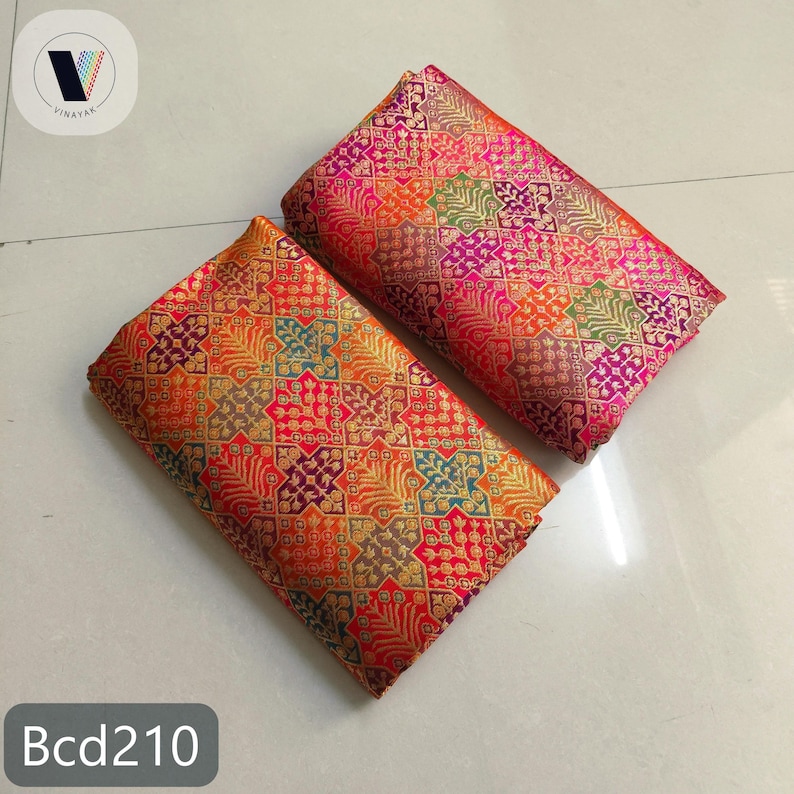 Puede incluir: Dos piezas de tela dobladas con intrincados patrones geom&eacute;tricos en tonos naranja, rojo, dorado, morado y verde. La tela tiene un aspecto brillante y texturizado. El texto "Bcd210" es visible en la parte inferior.