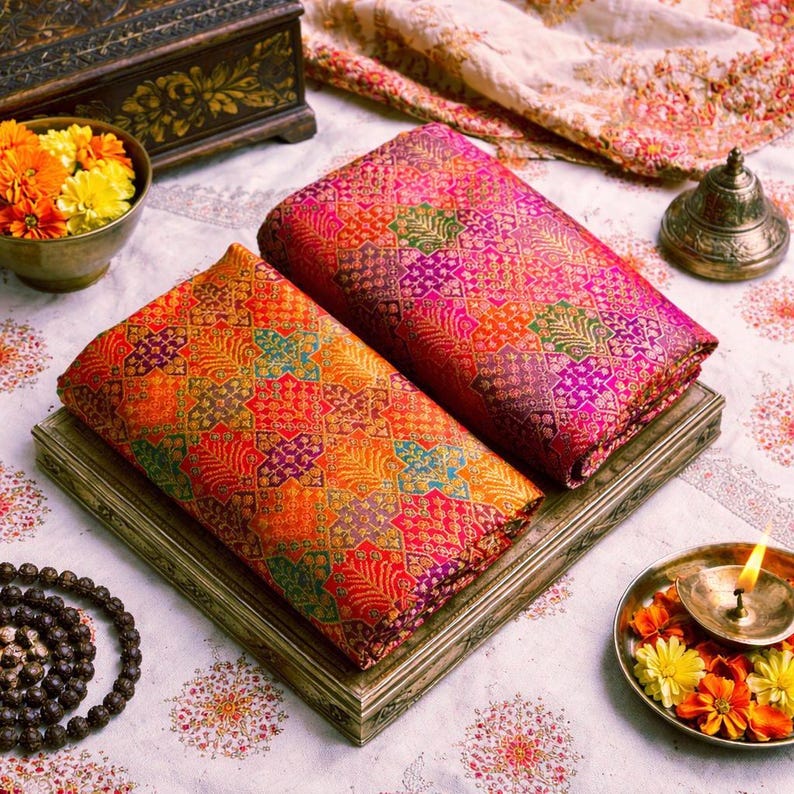 Puede incluir: Dos piezas de tela dobladas con un vibrante patr&oacute;n geom&eacute;trico repetitivo en tonos naranja, rosa, morado y verde. Los textiles est&aacute;n apilados sobre una bandeja rectangular, acompa&ntilde;ados por un cuenco de flores amarillas y naranjas.