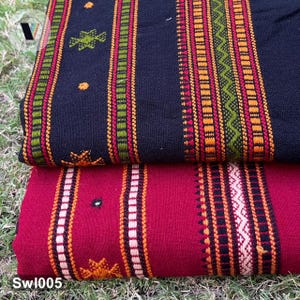 Puede incluir: Dos piezas de tela dobladas con patrones tejidos. La tela superior es negra con rayas verticales en verde, naranja y rojo, acentuadas por pequeños motivos bordados. La tela inferior es de color rojo intenso con rayas negras, blancas y naranjas.