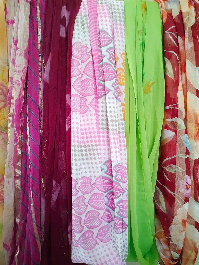 May include: A collection of vertical fabric scarves featuring diverse patterns and colours, including a pink tie-dye, a solid deep maroon, a white scarf with pink leaves and geometric shapes, a solid lime green, and a red floral print.