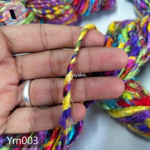 May include: Close-up of a strand of colorful yarn, held in a hand. The yarn is a mix of yellow, purple, green, red, and blue. The text "Yrn003" is visible at the bottom. The yarn is twisted and appears to be made of multiple strands.