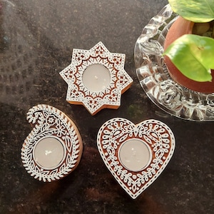 May include: Three wooden candle holders with white floral designs. The shapes include a star, a paisley, and a heart. Each holder has a circular space for a tea light candle. The candle holders are brown with white accents.