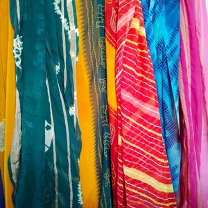 May include: A collection of long, rectangular scarves hanging vertically. The scarves feature various patterns and colours, including solid yellow, teal with white tie-dye stripes, yellow with green bandhani dots, red and yellow stripes, and blue tie-dye.