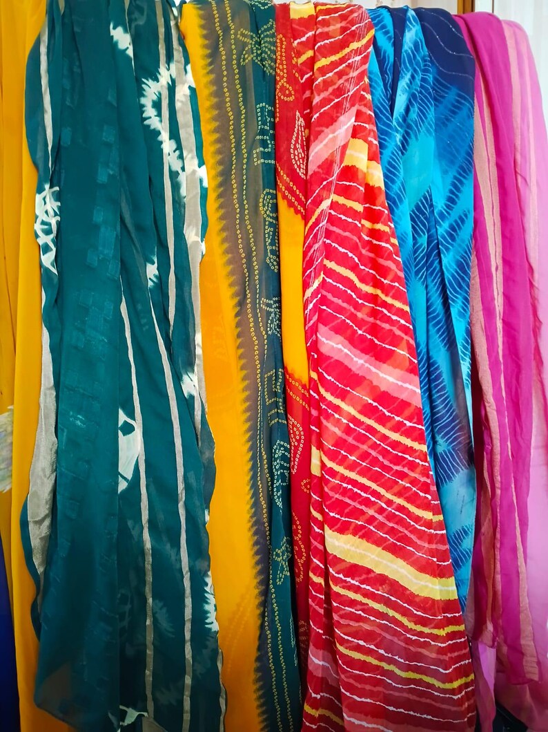 May include: A collection of long, rectangular scarves hanging vertically. The scarves feature various patterns and colours, including solid yellow, teal with white tie-dye stripes, yellow with green bandhani dots, red and yellow stripes, and blue tie-dye.