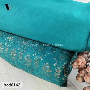 May include: Two fabric samples in shades of teal. The top fabric is a solid teal, while the bottom fabric features a teal background with a gold floral pattern. The text "Vinayak Textile India" is visible. The product code "bcd0142" is also visible.