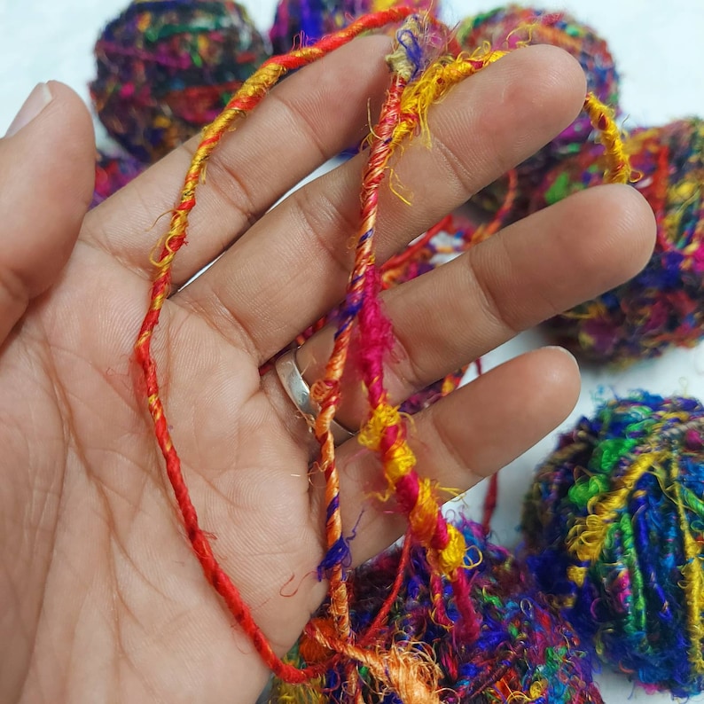 K&ouml;nnte beinhalten: Eine Hand h&auml;lt ein B&uuml;ndel bunter Wolle mit einem Farbschema aus Rot, Orange, Gelb, Gr&uuml;n, Blau und Lila. Die Wolle besteht aus verschiedenen Texturen, darunter glatt, flauschig und ausgefranst.