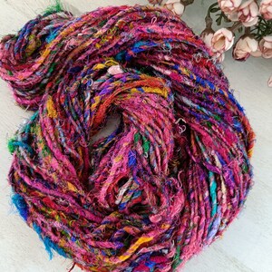K&ouml;nnte beinhalten: Ein Kn&auml;uel buntes, handgesponnenes Seiden-Garn mit einem Mix aus rosa, blauen, gr&uuml;nen und gelben F&auml;den. Das Garn wird aus recycelten Saris hergestellt und ist perfekt zum Stricken oder H&auml;keln.