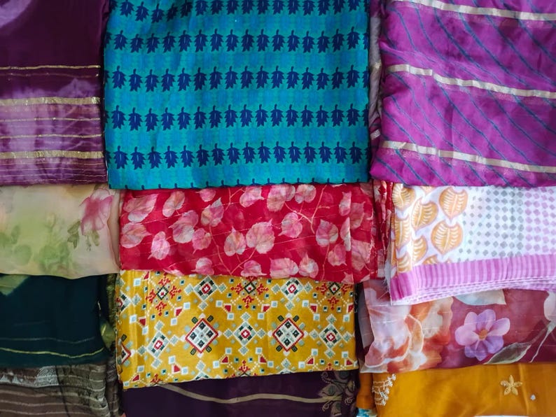 May include: A collection of folded fabric pieces featuring diverse patterns and colours. Visible designs include a teal leaf print, a red leaf pattern, a yellow geometric motif, and various floral prints in shades of purple, pink, and gold.