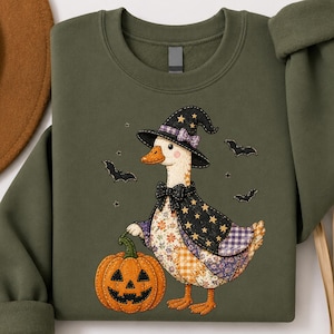 May include: A military green crewneck sweatshirt featuring a graphic of a duck wearing a witch hat and a bow tie. The duck is standing next to a jack-o'-lantern pumpkin, surrounded by small black bats and stars on a patterned patchwork body.