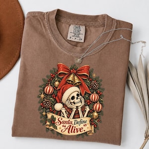 Könnte beinhalten: Ein braunes T-Shirt mit einer Grafik eines Skeletts mit Weihnachtsmütze, zentriert in einem Weihnachtskranz mit roten Kugeln, Tannenzapfen und einer großen roten Schleife. Unter dem Skelett befindet sich ein Banner mit dem Text Santa, Define Alive.