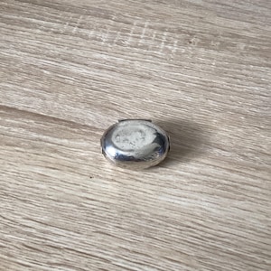 May include: A small, oval-shaped silver pill box with a polished, reflective metallic surface. The container features a hinged lid design with a smooth, rounded top and visible seams along the sides, showcasing a simple and compact construction.