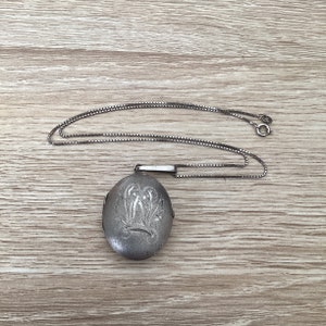 Può includere: Un ciondolo a medaglione ovale in argento con un motivo floreale inciso sulla superficie anteriore. Il medaglione è fissato a una sottile e delicata catenina d'argento a maglie quadrate, dotata di una piccola chiusura a molla circolare.