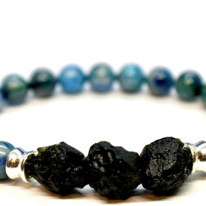 Op de afbeelding: Een armband met blauwe en groene bolvormige kralen. De armband heeft drie grote, ruwe zwarte stenen in het midden, gescheiden door zilverkleurige metalen kralen. De armband staat op een witte achtergrond.