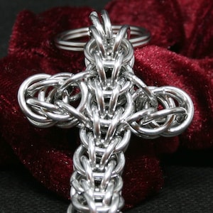 Pode incluir: Um chaveiro de metal prateado em forma de cruz, feito de anéis entrelaçados. A cruz está sobre um fundo de veludo vermelho.