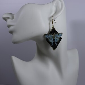 May include: A dangling earring crafted from small beads in a geometric moth shape. The design features a light blue body and wings contrasted against a black background, all outlined with gold-toned beads and suspended from a gold-toned metal hook.