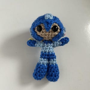 May include: A handmade crochet doll of a video game character. The doll features a blue helmet, body, and boots, with a tan face and black eyes. The arms are outstretched, and the doll is approximately 4 inches tall.