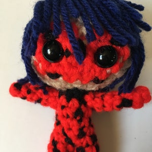 May include: A crocheted plush toy of a superhero with red and black patterned clothing, blue hair, and black eyes.