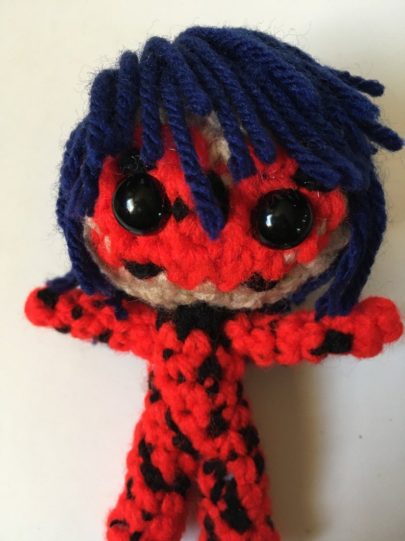 May include: A crocheted plush toy of a superhero with red and black patterned clothing, blue hair, and black eyes.