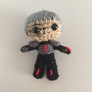 May include: A handmade crochet doll with a gray head of hair, tan face, and large black eyes. The doll wears a black outfit with gray accents and red details. The doll is standing upright with arms outstretched.