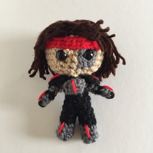 May include: A handmade crochet doll with a warrior theme. The doll has a brown yarn hairstyle, a red headband, and large black eyes. The doll's outfit is black with gray and red accents. The doll is approximately 6 inches tall.