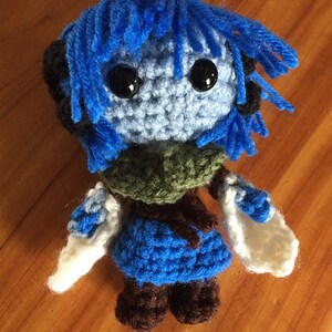 May include: A blue crocheted plush toy with black eyes and brown boots. The toy has blue hair and is wearing a green and blue outfit.