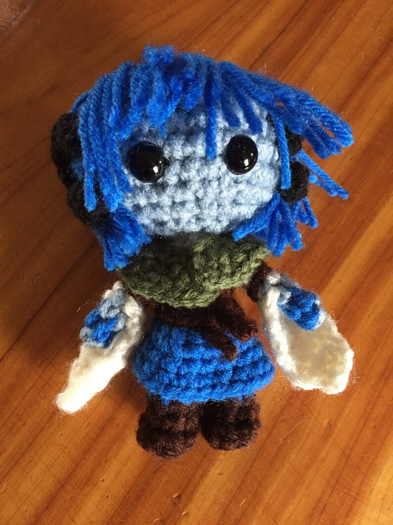 May include: A blue crocheted plush toy with black eyes and brown boots. The toy has blue hair and is wearing a green and blue outfit.