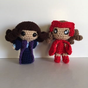 May include: Two crocheted dolls, one in a purple robe and the other in a red cape and dress. Both dolls have brown hair and large black eyes.