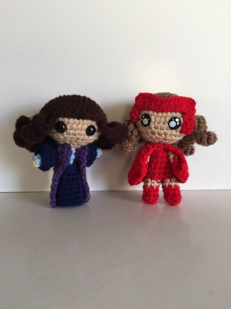 May include: Two crocheted dolls, one in a purple robe and the other in a red cape and dress. Both dolls have brown hair and large black eyes.