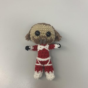 May include: A handmade crochet doll with a dog-like head and a red and white outfit. The doll has black button eyes, brown yarn hair, and a small, dark brown muzzle. The outfit includes a red top and pants with white accents.