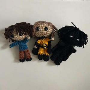 May include: Three handmade crochet dolls. One doll has brown hair and a blue shirt, another has tan hair and a yellow outfit, and the third is all black. Each doll has black eyes and is made of yarn.