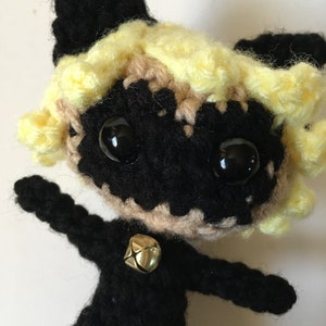 May include: Crocheted plush toy of a black cat with yellow hair, wearing a black suit and a gold bell. The cat has large black eyes and a black mask.