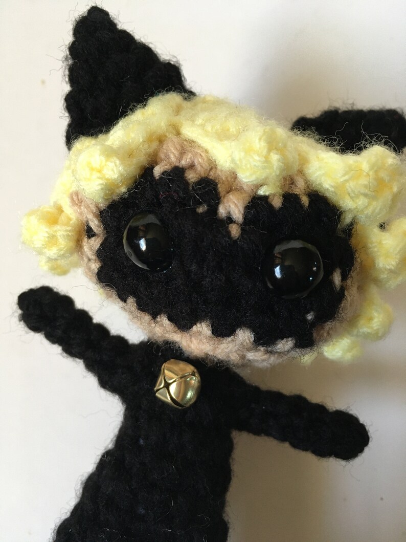 May include: Crocheted plush toy of a black cat with yellow hair, wearing a black suit and a gold bell. The cat has large black eyes and a black mask.