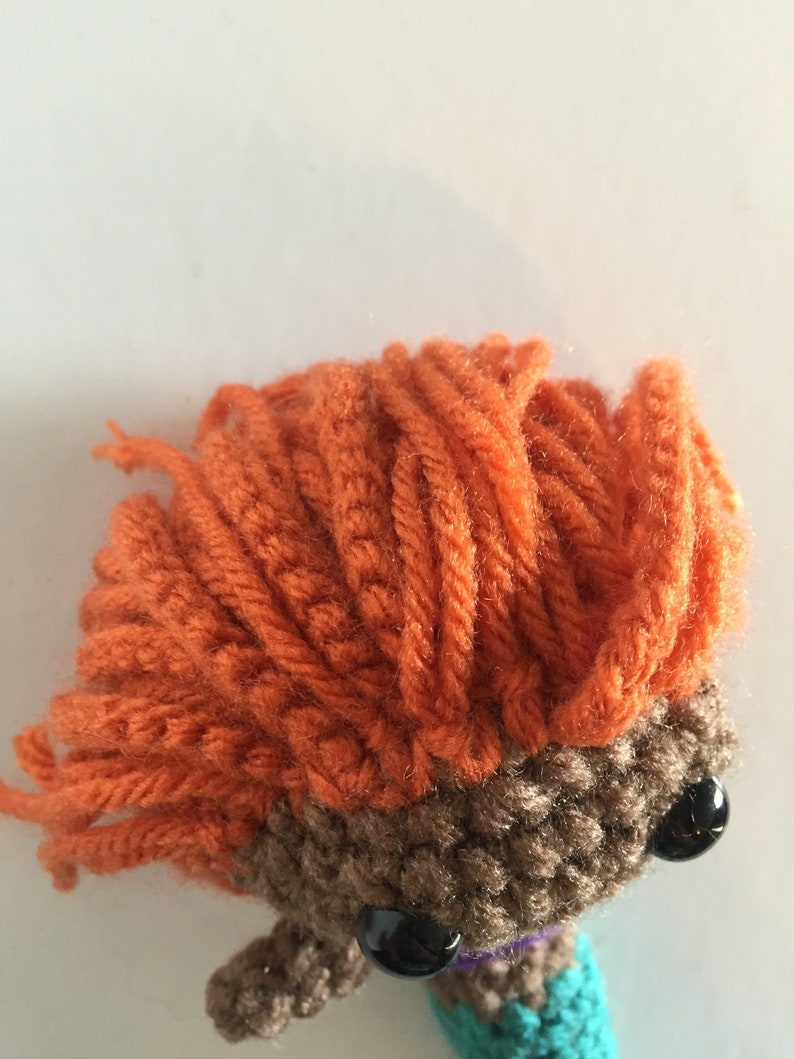 May include: A crocheted mermaid doll with brown skin, orange hair, and black eyes. The doll is wearing a turquoise and purple tail.