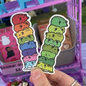 May include: Two stacks of rainbow frog stickers. Each stack has six frogs, each a different color of the rainbow. The frogs are all smiling and have big eyes.