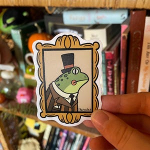 May include: A sticker of a frog wearing a top hat and a suit, in a gold frame. The frog is green with a white shirt and a black tie. The sticker has the text "thcreates" in the bottom right corner.