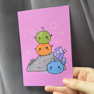 May include: A pink card with a cartoon illustration of three round characters stacked on top of each other. The top character is green, the middle character is orange, and the bottom character is blue. The characters are on a gray rock with purple crystals and mushrooms. The card has a pink background with white stars.
