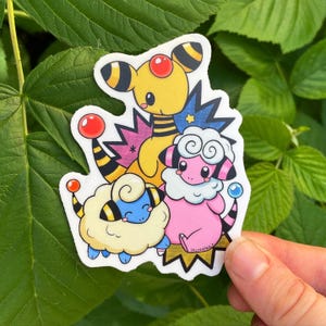 May include: A colorful sticker featuring three cartoon characters. The characters are yellow, pink, and blue, with black and white striped accents. The sticker is held against a background of green leaves.